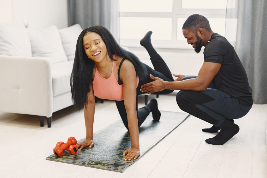 Man and woman exercising on mats in a bright living room using weights and stretching exercises.